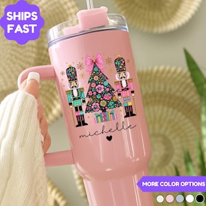 May include: Pink insulated tumbler with a handle, featuring a Christmas tree and nutcracker design. The tumbler has a clear lid and a straw. The name "michelle" is written on the tumbler. The words "Ships Fast" are in a purple circle.