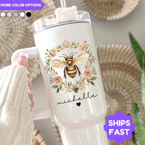 May include: White insulated tumbler with a bee and floral design. The tumbler features a watercolor-style bee illustration surrounded by a wreath of pink flowers and green leaves. The name "michelle" is written in script below the bee, with a small black heart. The tumbler has a white handle and lid.