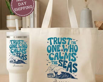 Könnte beinhalten: Eine weiße Stofftasche und ein passender weißer Becher mit Strohhalm. Beide Artikel zeigen den blauen Schriftzug Trust the One Who Calms the Seas, eine Wellengrafik, den Text Mark 4:35-41 sowie den Namen Jessica in schwarzer Schreibschrift.