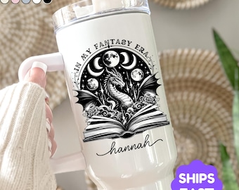 May include: White insulated tumbler with a black and white fantasy-themed design. The design includes a dragon, moons, and the text "IN MY FANTASY ERA" and "hannah". The tumbler has a handle and a clear lid.