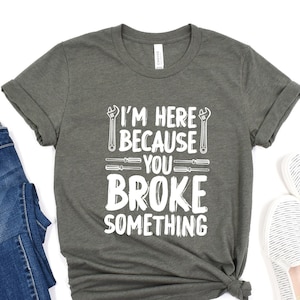 May include: A dark gray t-shirt with the text "I'm here because you broke something" in white. The text is surrounded by images of tools, including a wrench and a screwdriver.