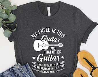 May include: A dark gray t-shirt with a white graphic that says "All I need is this Guitar and that other Guitar" with a drawing of a guitar. The text continues with "and those guitars over there and the guitars in the other rooms, and..."