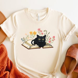 Puede incluir: Una camiseta blanca con un gato negro sentado en un libro abierto rodeado de flores de colores. El gato mira directamente al espectador.