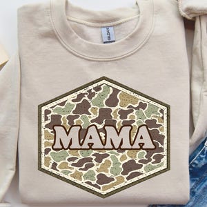 May include: Beige sweatshirt with a camouflage hexagon design that reads "MAMA" in a bold, neutral font. The sweatshirt is laid flat with rolled-up sleeves, next to a pair of blue jeans and a coffee cup.