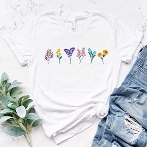 May include: White t-shirt with a row of colourful flowers printed on the front. The shirt is lying flat on a white marble surface with a pair of blue jeans and a sprig of greenery.