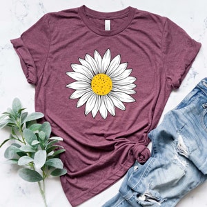 May include: A maroon t-shirt with a white daisy graphic. The daisy has a yellow center and is centered on the shirt. The shirt is laying on a white marble surface with a pair of blue jeans and a sprig of green foliage.