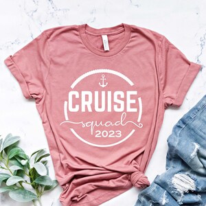 May include: A pink t-shirt with a white circle design that says "Cruise Squad 2023" with an anchor symbol. The shirt is laying on a white marble surface with a pair of blue jeans.