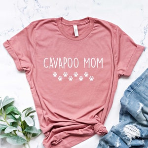 May include: A dusty rose t-shirt with the text "CAVAPOO MOM" in white lettering and a row of white paw prints below the text. The t-shirt is laying on a white marble surface with a pair of blue jeans with distressed details.