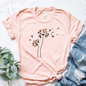 May include: A light pink t-shirt with a graphic of two dandelion flowers with butterflies on them. The butterflies are in shades of brown, green, and orange.