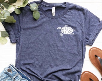 May include: A navy blue t-shirt with a white sea turtle graphic. The shirt is laid flat on a white surface with a pair of brown sandals and a pair of blue denim shorts.