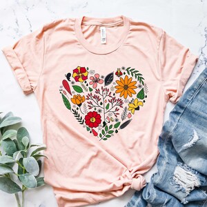 May include: A light pink t-shirt with a floral heart design. The heart is made up of various flowers, leaves, and a bee. The shirt is laid flat on a white marble surface.