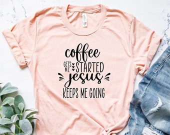 May include: A peach-colored t-shirt with black text that reads "coffee gets me started jesus keeps me going". The shirt is laid out on a white marble surface with a pair of blue jeans.