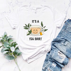 May include: White t-shirt with a graphic of a teacup with a smiling face and the text "It's a Tea Shirt".