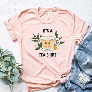 May include: A pink t-shirt with the text "It's a Tea Shirt" and a cartoon illustration of a teacup with a smiling face.