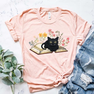 Puede incluir: Una camiseta rosa con un gato negro acostado sobre un libro abierto. El libro est&aacute; rodeado de flores de colores.