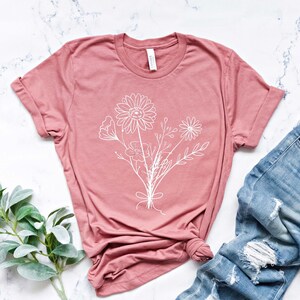 Pode incluir: Uma camiseta rosa p&oacute; com um contorno branco de um buqu&ecirc; de flores. A camiseta est&aacute; sobre uma superf&iacute;cie de m&aacute;rmore branco com um par de jeans azul claro desbotados.
