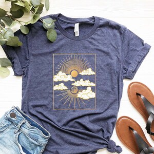 May include: A navy blue t-shirt with a gold sun and moon graphic on a white cloud background. The graphic is framed in a gold rectangle.