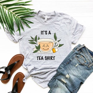 May include: A gray t-shirt with the text "It's a Tea Shirt" and a cartoon image of a smiling teacup with a tea bag.