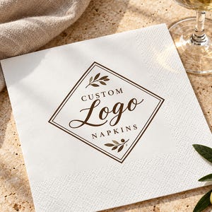 Może przedstawiać: Kwadratowa biała serwetka papierowa z brązową obwódką w kształcie rombu oraz motywem liści u góry i u dołu. Napis CUSTOM Logo NAPKINS jest nadrukowany na środku przy użyciu kombinacji czcionek szeryfowych i skryptowych w kolorze ciemnobrązowym.