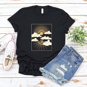 May include: A black t-shirt with a gold graphic of a sun, moon, and clouds. The graphic is in a rectangular frame.