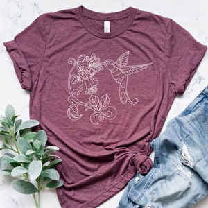 May include: A maroon t-shirt with a white outline of a hummingbird drinking nectar from a flower.