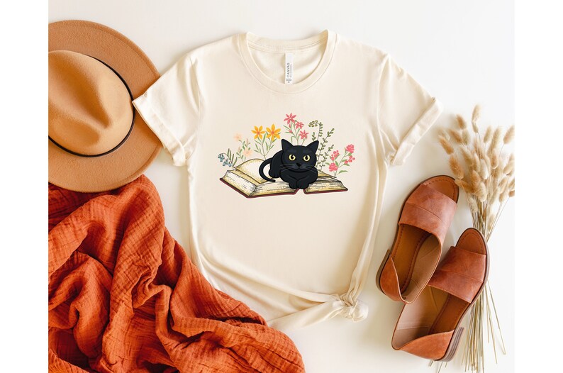 Puede incluir: Una camiseta blanca con un gato negro sentado en un libro abierto rodeado de flores de colores. El gato mira directamente al espectador.