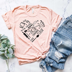 Może przedstawiać: R&oacute;żowy t-shirt z czarnym nadrukiem w kształcie serca, przedstawiającym składniki i narzędzia do pieczenia. Tekst na nadruku zawiera słowa: "s&oacute;l", "mleko", "wanilia", "kakao", "śmietanka", "cukier", "jajka", "cynamon" i "mąka".