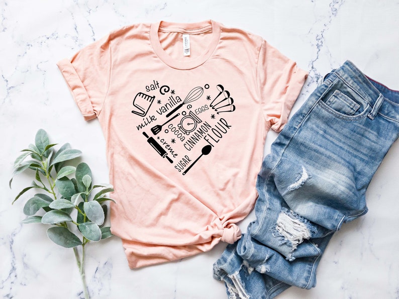 Może przedstawiać: R&oacute;żowy t-shirt z czarnym nadrukiem w kształcie serca, przedstawiającym składniki i narzędzia do pieczenia. Tekst na nadruku zawiera słowa: "s&oacute;l", "mleko", "wanilia", "kakao", "śmietanka", "cukier", "jajka", "cynamon" i "mąka".