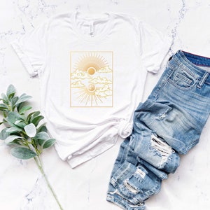May include: White t-shirt with a gold graphic of a sun and moon with clouds in a square frame.