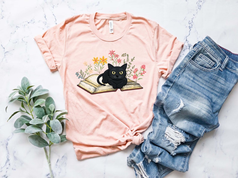 Puede incluir: Una camiseta rosa con un gato negro acostado sobre un libro abierto. El libro est&aacute; rodeado de flores de colores.