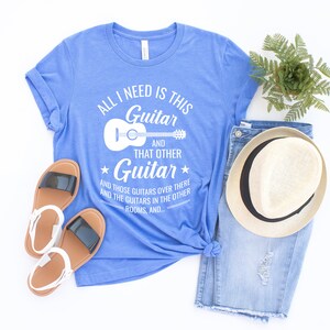 Puede incluir: Una camiseta azul con texto blanco que dice "All I need is this Guitar and that other Guitar and those guitars over there and the guitars in the other rooms, and..." con un contorno blanco de una guitarra ac&uacute;stica.