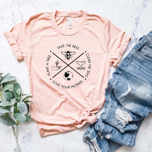 May include: A light pink t-shirt with a circular graphic featuring a bee, whale tail, globe, and hand planting a tree. The text reads "Save the Bees", "Clean the Seas", "Love Your Mother", and "Plant a Tree". Blue denim jeans are next to the shirt.