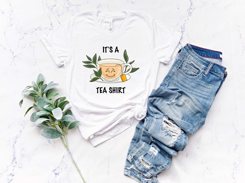May include: White t-shirt with a graphic of a teacup with a smiling face and the text "It's a Tea Shirt".