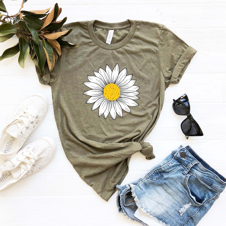 May include: A green t-shirt with a white daisy graphic. The daisy has a yellow center. The t-shirt is laying on a white surface with a pair of white shoes, a pair of denim shorts, and sunglasses.