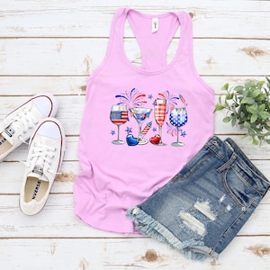 Pode incluir: Camisola de al&ccedil;as rosa com um gr&aacute;fico de quatro copos com desenhos vermelho, branco e azul, fogos de artif&iacute;cio e estrelas. Os copos est&atilde;o cheios de diferentes bebidas. A camisola est&aacute; sobre uma superf&iacute;cie de madeira branca com um par de t&eacute;nis brancos e cal&ccedil;&otilde;es de ganga azuis.