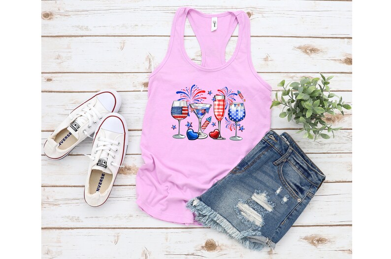 Pode incluir: Camisola de al&ccedil;as rosa com um gr&aacute;fico de quatro copos com desenhos vermelho, branco e azul, fogos de artif&iacute;cio e estrelas. Os copos est&atilde;o cheios de diferentes bebidas. A camisola est&aacute; sobre uma superf&iacute;cie de madeira branca com um par de t&eacute;nis brancos e cal&ccedil;&otilde;es de ganga azuis.