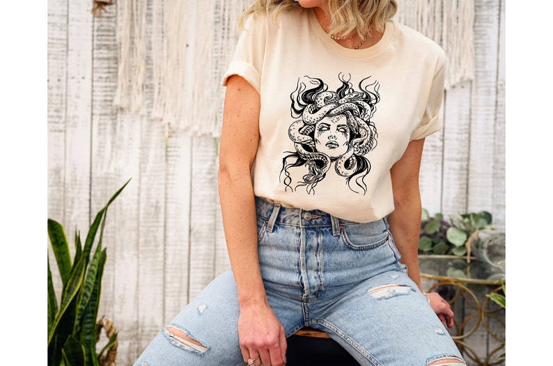 Pode incluir: Uma camiseta creme com uma ilustra&ccedil;&atilde;o em preto e branco do rosto da Medusa. O gr&aacute;fico mostra Medusa com cobras no cabelo. A modelo est&aacute; usando jeans azul claro.