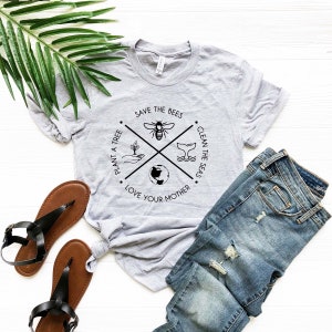 May include: A heather gray t-shirt with a circular graphic that reads "Save the Bees," "Clean the Seas," "Plant a Tree," and "Love Your Mother." The shirt is paired with blue jeans and brown sandals. A green leaf is in the background.