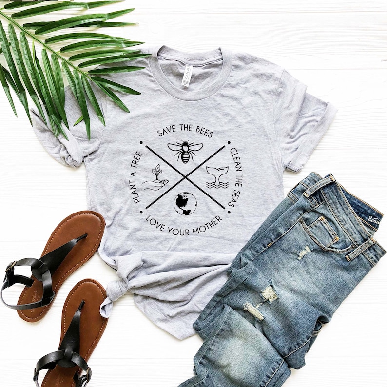 May include: A heather gray t-shirt with a circular graphic that reads "Save the Bees," "Clean the Seas," "Plant a Tree," and "Love Your Mother." The shirt is paired with blue jeans and brown sandals. A green leaf is in the background.