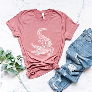 Puede incluir: Una camiseta rosa con un gr&aacute;fico de cocodrilo blanco. La camiseta est&aacute; colocada sobre una superficie de m&aacute;rmol blanco con un par de pantalones vaqueros azules y una rama de vegetaci&oacute;n.