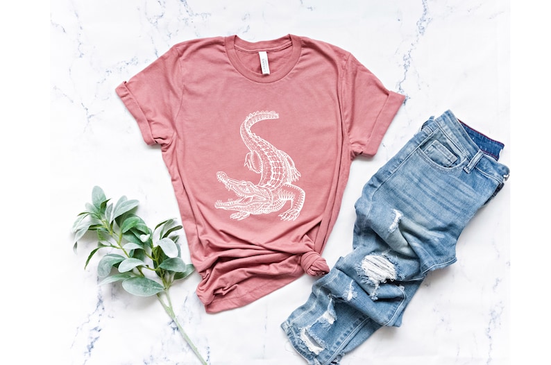 Puede incluir: Una camiseta rosa con un gr&aacute;fico de cocodrilo blanco. La camiseta est&aacute; colocada sobre una superficie de m&aacute;rmol blanco con un par de pantalones vaqueros azules y una rama de vegetaci&oacute;n.