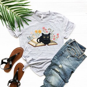 Puede incluir: Una camiseta gris con un gato negro sentado en un libro abierto rodeado de flores de colores.