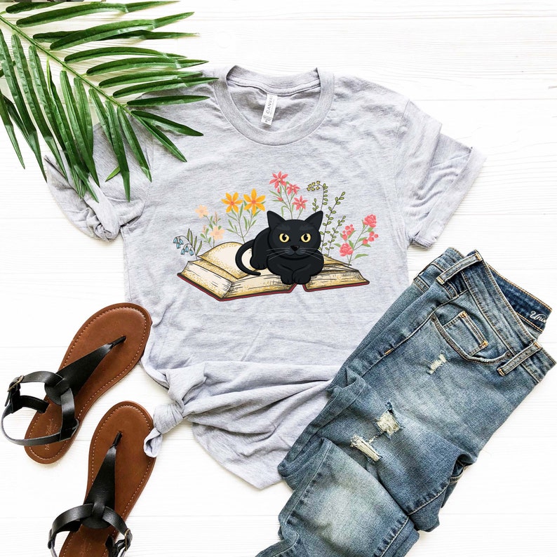 Puede incluir: Una camiseta gris con un gato negro sentado en un libro abierto rodeado de flores de colores.