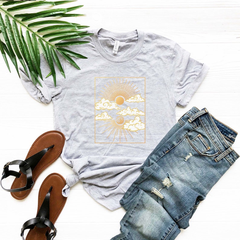May include: A gray t-shirt with a gold sun and moon graphic design. The design features a sun with rays and a crescent moon with stars, both surrounded by clouds. The graphic is framed in a gold rectangle.