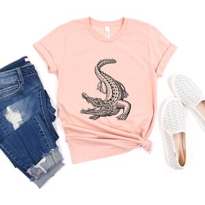 Puede incluir: Una camiseta rosa con una ilustraci&oacute;n en blanco y negro de un cocodrilo en la parte delantera. La camiseta se combina con unos vaqueros azules y unas zapatillas blancas de deslizamiento.