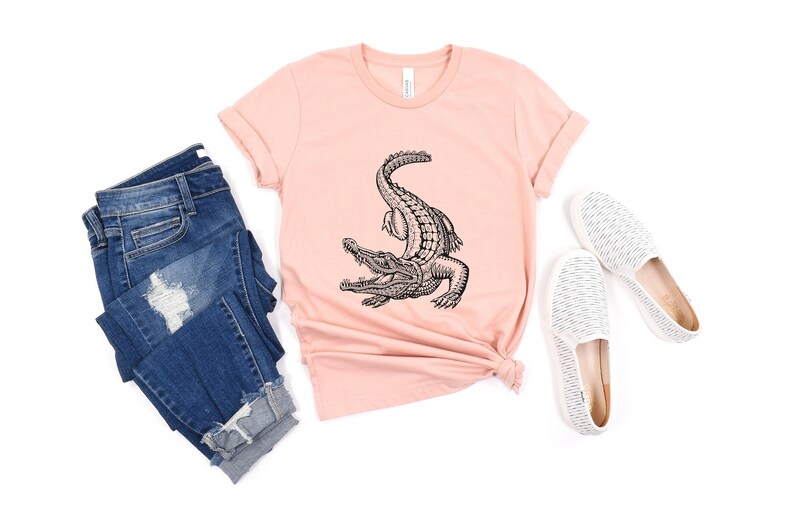 Puede incluir: Una camiseta rosa con una ilustraci&oacute;n en blanco y negro de un cocodrilo en la parte delantera. La camiseta se combina con unos vaqueros azules y unas zapatillas blancas de deslizamiento.