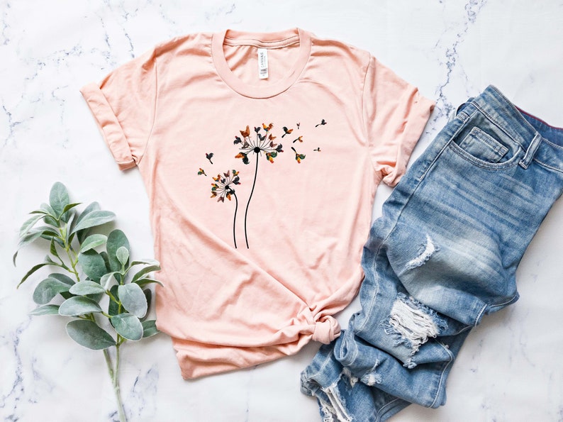 May include: A light pink t-shirt with a graphic of two dandelion flowers with butterflies on them. The butterflies are in shades of brown, green, and orange.