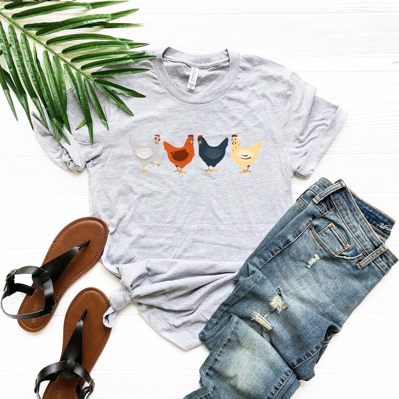 Może przedstawiać: Szary t-shirt z nadrukiem czterech kurczak&oacute;w w rzędzie. Kurczaki są białe, brązowe, czarne i ż&oacute;łte.