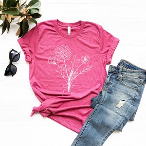 Pode incluir: Uma camiseta rosa com um contorno branco de um buqu&ecirc; de flores. A camiseta est&aacute; colocada sobre uma superf&iacute;cie branca com um par de cal&ccedil;as de ganga azuis.