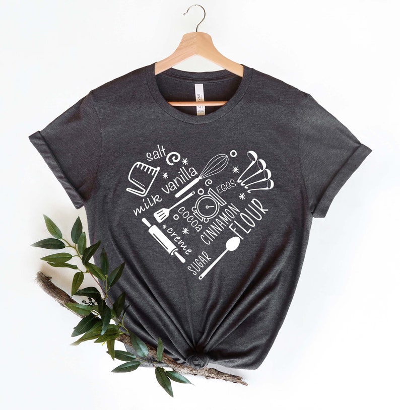Może przedstawiać: Szary t-shirt z białym nadrukiem przedstawiającym kuchenne narzędzia i składniki do pieczenia. Tekst na koszulce brzmi: "s&oacute;l", "mleko", "wanilia", "śmietanka", "cukier", "kakao", "jajka", "cynamon" i "mąka".